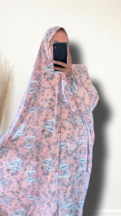 A person taking a mirror selfie wearing a light pink modest garment with a blue and gray floral print, holding a black smartphone in front of their face against a plain off-white wall.