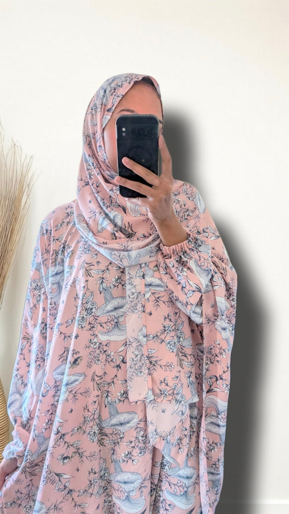 A woman takes a mirror selfie wearing a light pink hijab and a matching long-sleeve kaftan-style dress with a blue and gray floral print, set against a plain off-white wall.