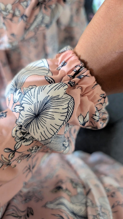 A close-up of a person's wrist wearing a sheer, gathered fabric wristlet or cuff in a dusty pink color with a black floral and botanical line-art print.