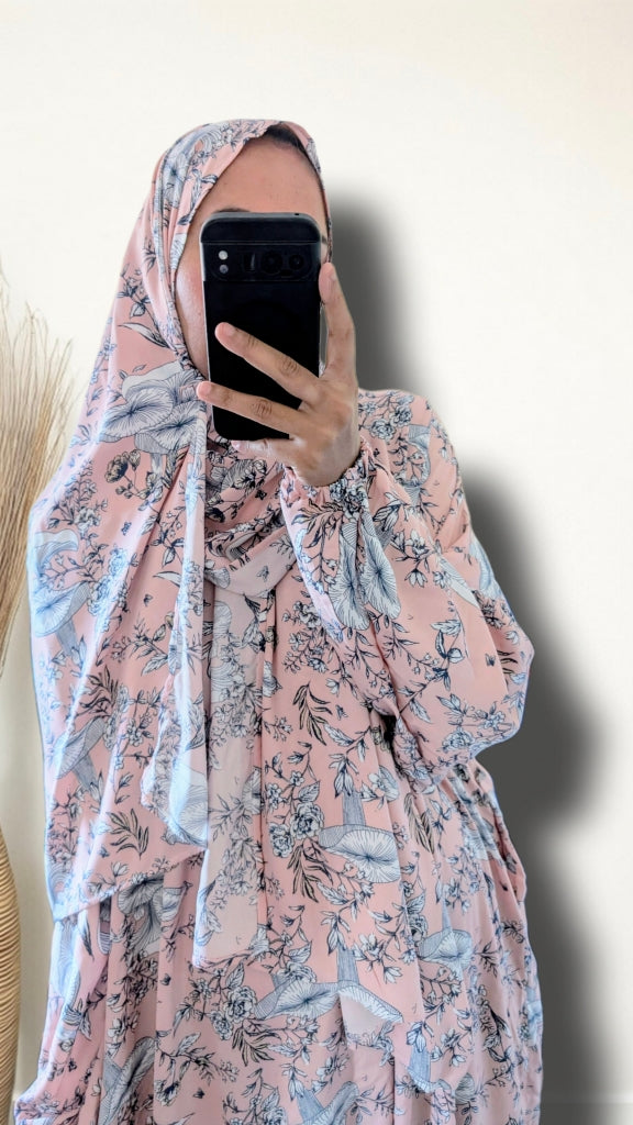 A woman takes a mirror selfie wearing a light pink modest dress with a blue and gray floral print, paired with a matching patterned hijab. She holds a black smartphone to block her face against a plain off-white wall.