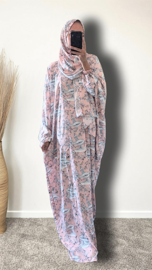 A woman wearing a long, loose-fitting modest dress with a pink and blue floral print, paired with a matching headscarf, taking a mirror selfie in an indoor setting.