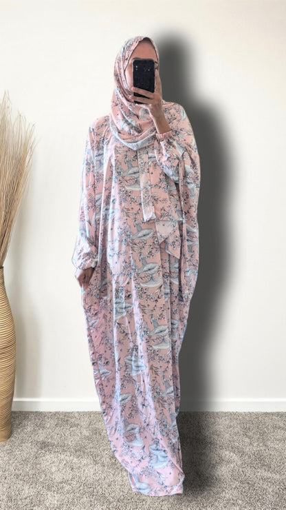 A woman wearing a long, loose-fitting modest dress with a pink and blue floral print, paired with a matching headscarf, taking a mirror selfie in an indoor setting.