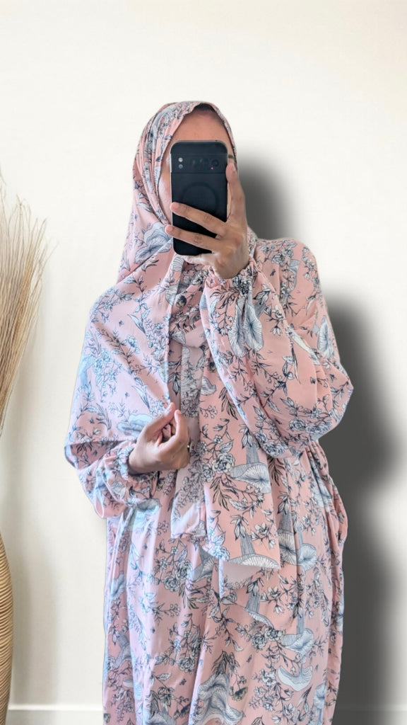 A woman takes a mirror selfie wearing a pink and blue floral print hijab and matching long-sleeve abaya dress. She is holding a black smartphone in front of her face, with a plain off-white wall behind her and a dried pampas grass decoration on the left.