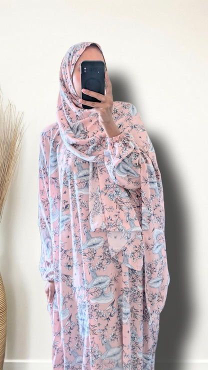 A woman wearing a pink and gray floral print hijab and matching long-sleeve abaya, taking a mirror selfie with a black smartphone against a plain beige wall.