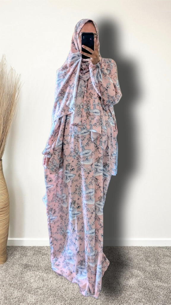 A woman wearing a full-length modest outfit with a matching headscarf, featuring a pink and light blue abstract pattern, taking a mirror selfie in an indoor setting.