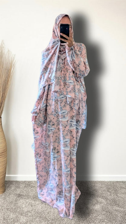 A woman wearing a full-length modest outfit with a matching headscarf, featuring a pink and light blue abstract pattern, taking a mirror selfie in an indoor setting.