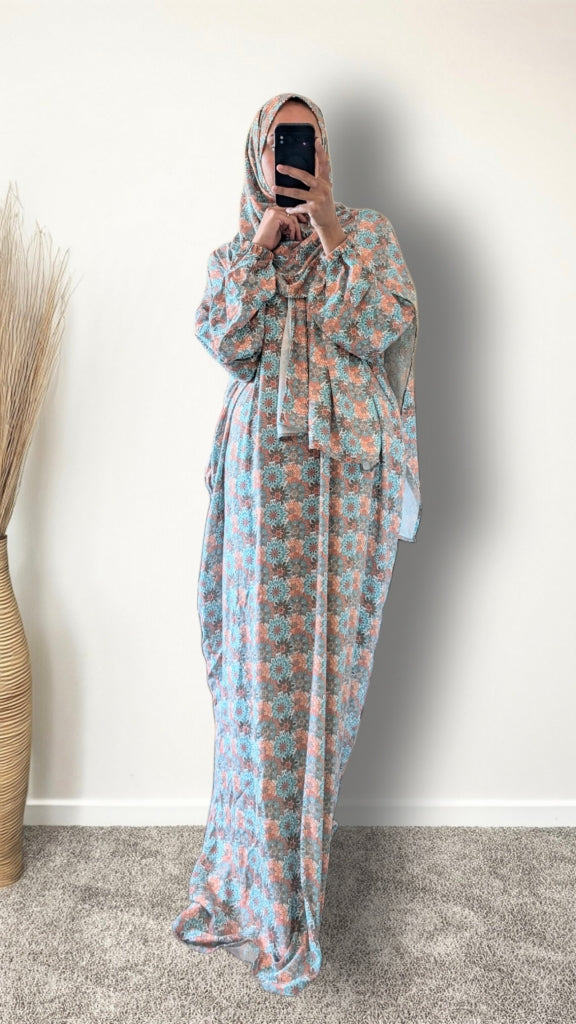 A woman wearing a long, loose-fitting maxi abaya dress with a light blue and peach floral pattern, paired with a matching hijab, taking a mirror selfie in an indoor setting.