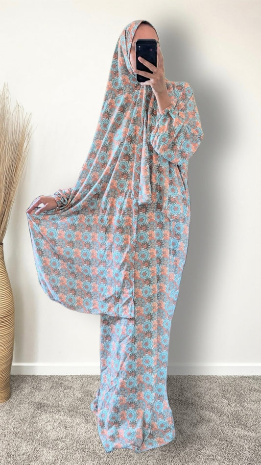 A woman takes a mirror selfie wearing a full-length modest outfit with a matching head covering. The garment has a light blue base with a repeating coral-pink floral pattern and is styled in a home setting with a beige wall and a woven vase on the left.