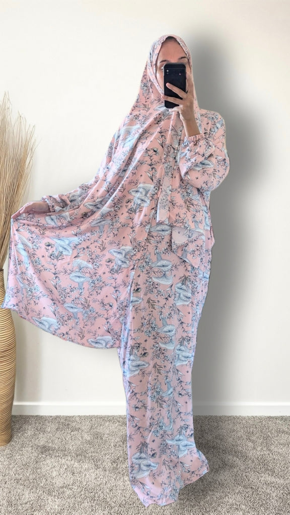A woman takes a mirror selfie wearing a full-body pink and blue floral print shapter or modest robe with a matching head covering, standing indoors against a plain wall.