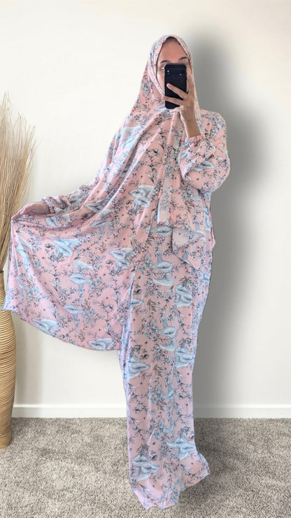A woman takes a mirror selfie wearing a full-body pink and blue floral print shapter or modest robe with a matching head covering, standing indoors against a plain wall.