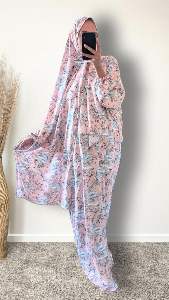 A person takes a mirror selfie wearing a full-body pink and blue patterned garment with a matching head covering, standing indoors against a plain wall on a carpeted floor.